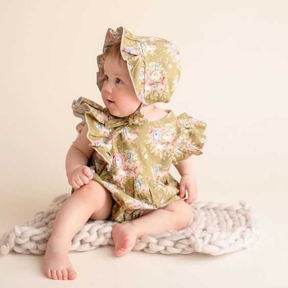 Baby Wearing Mustard Floral Ruffle Romper and Ruffle Bonnet White Sitting on Chunky Knit Blanket